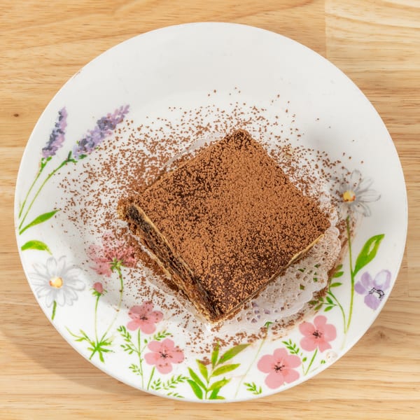 A square slice of tiramisu, dusted with cocoa powder, on a floral plate.