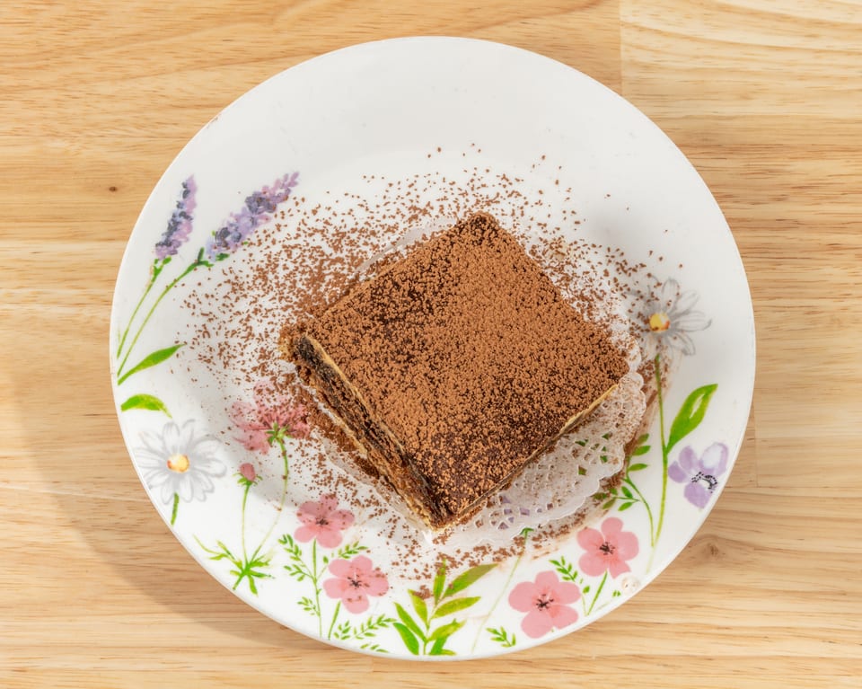 A square slice of tiramisu, dusted with cocoa powder, on a floral plate.