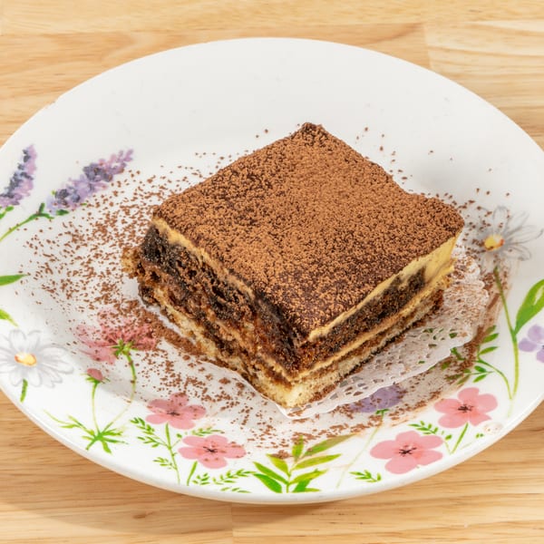 A slice of tiramisu with cocoa powder on a floral white plate, on a wooden table.