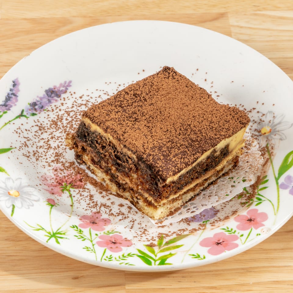 A slice of tiramisu with cocoa powder on a floral white plate, on a wooden table.