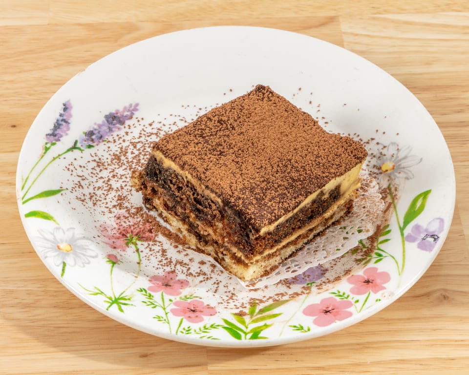 A slice of tiramisu with cocoa powder on a floral white plate, on a wooden table.