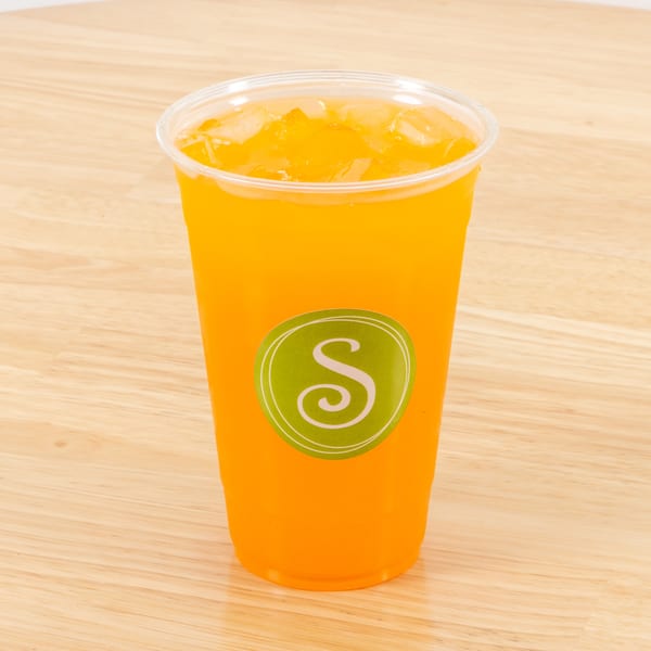 A clear plastic cup filled with orange juice and ice, featuring a green "S" logo, on a wooden table.