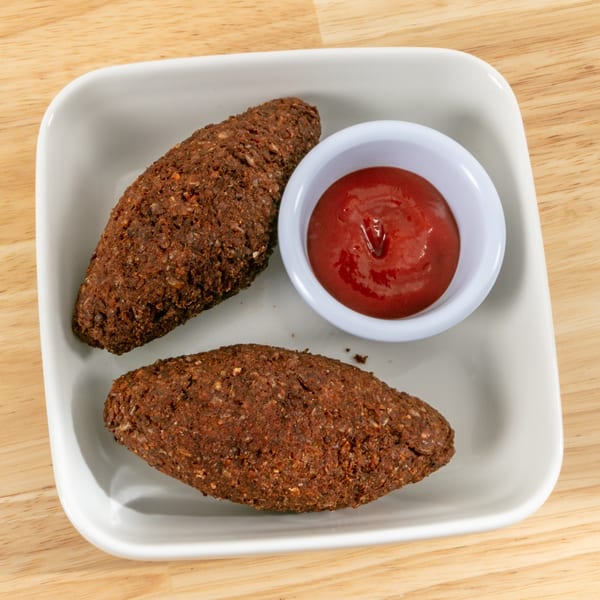 Two kibbeh, football-shaped fried meat and bulgur croquettes, with a side of ketchup on a white plate.