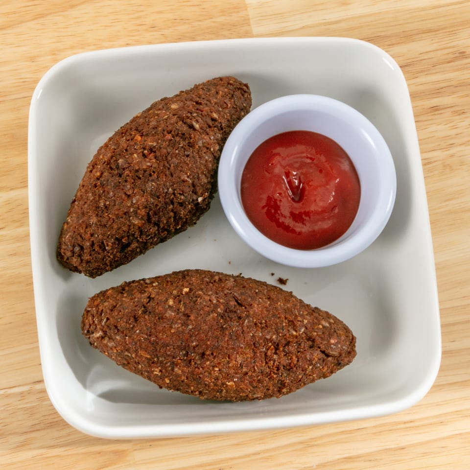 Two kibbeh, football-shaped fried meat and bulgur croquettes, with a side of ketchup on a white plate.