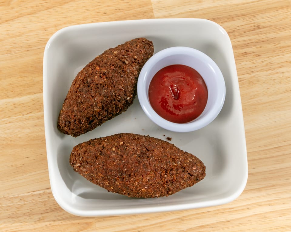 Two kibbeh, football-shaped fried meat and bulgur croquettes, with a side of ketchup on a white plate.
