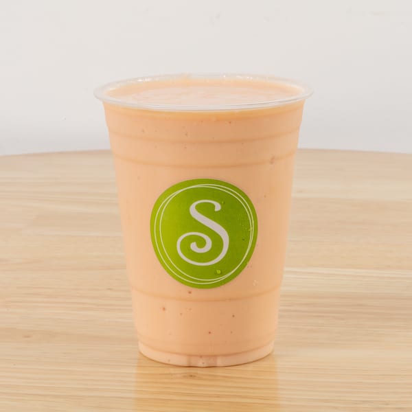 A peach-colored smoothie in a clear plastic cup with a green 'S' logo on a wooden table.
