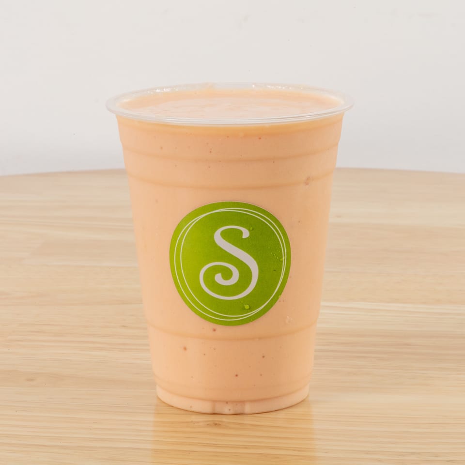 A peach-colored smoothie in a clear plastic cup with a green 'S' logo on a wooden table.