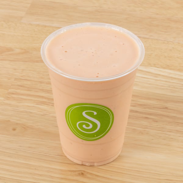 A peach-colored smoothie in a clear plastic cup with a green "S" logo on a wooden surface.