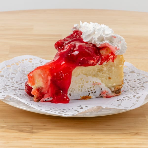 Slice of cheesecake with strawberry topping and whipped cream on a white doily.