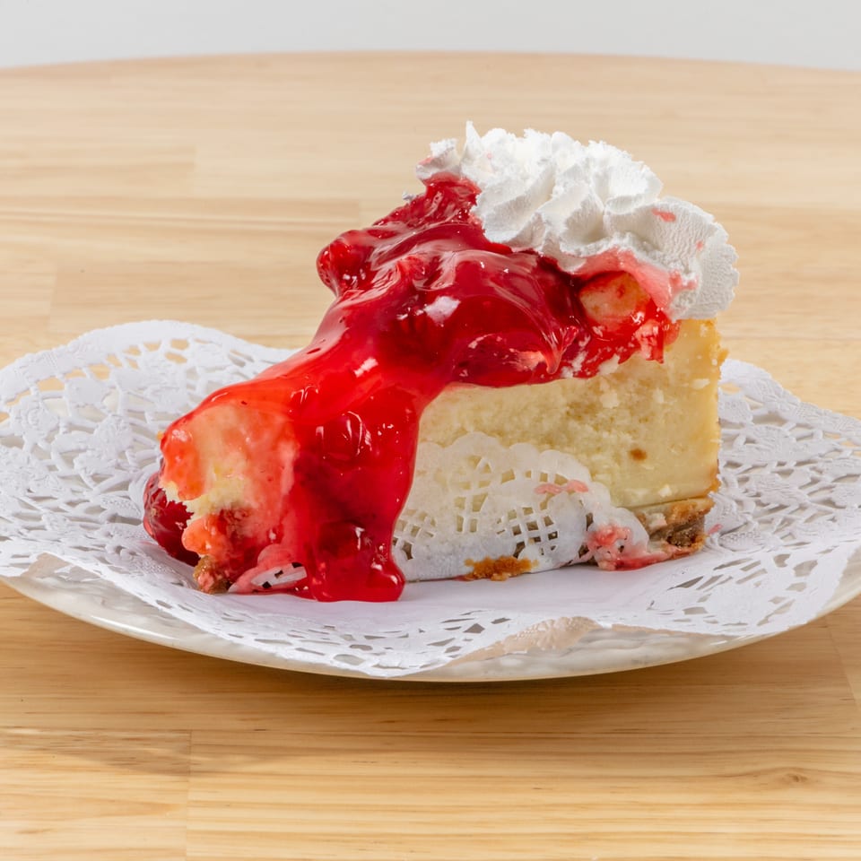 Slice of cheesecake topped with strawberry sauce and whipped cream on a white doily.