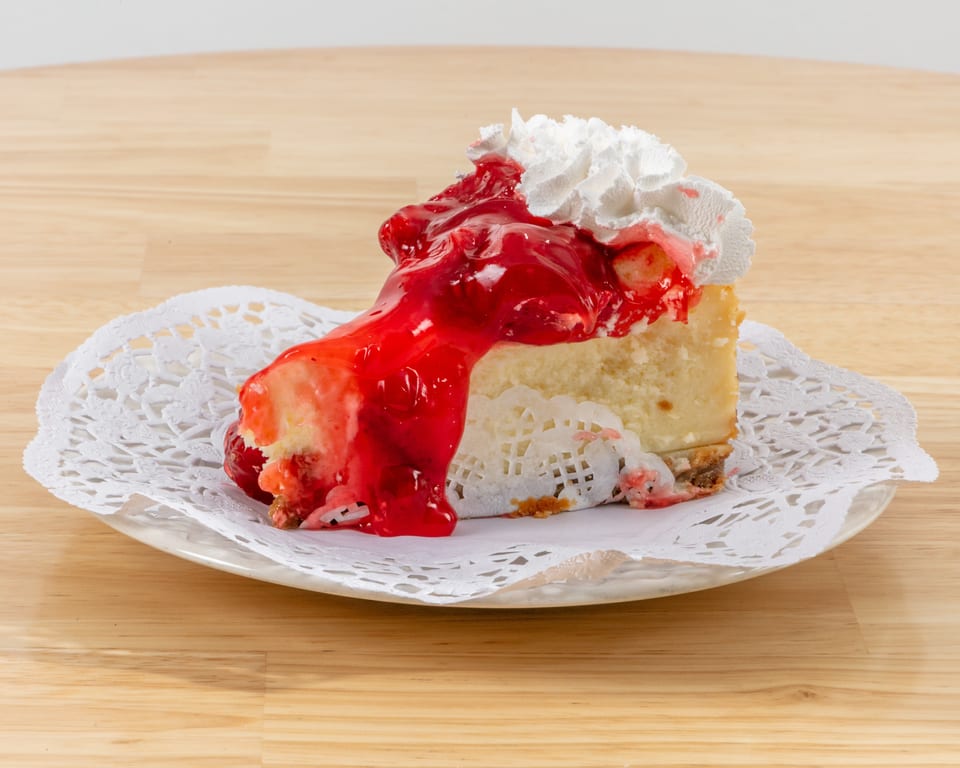 Slice of cheesecake topped with strawberry sauce and whipped cream on a white doily.