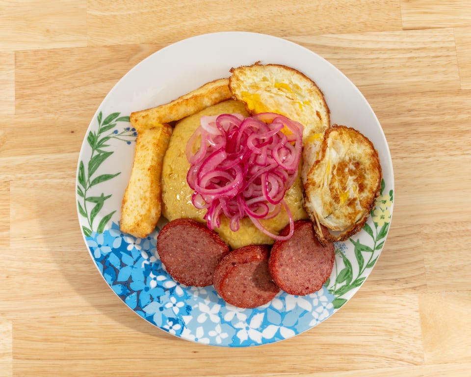 Mangu with fried eggs, salami, fried cheese, and pickled red onions on a floral plate.