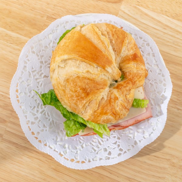 Ham and cheese croissant sandwich with lettuce on a white doily, on a wooden table.