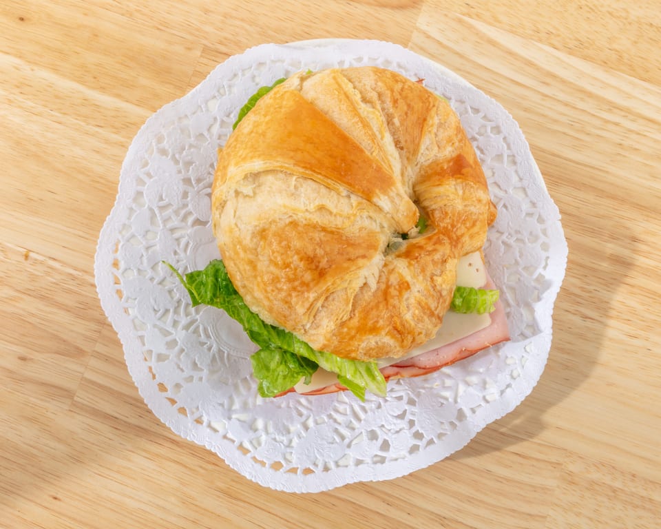 Ham and cheese croissant sandwich with lettuce on a white doily, on a wooden table.