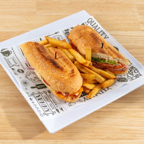Two submarine sandwiches with fries on a white plate with decorative paper on a wooden table.