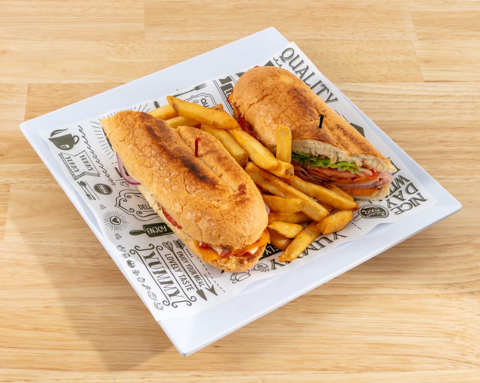 Two submarine sandwiches with fries on a white plate with decorative paper on a wooden table.