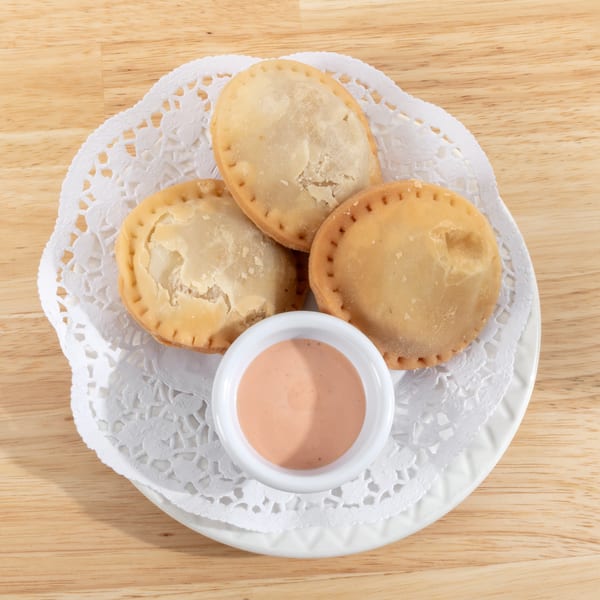 Three golden-brown empanadas and a small dish of pink dipping sauce on a white doily.