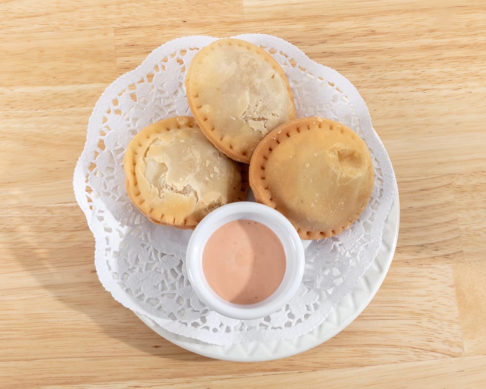 Three golden-brown empanadas and a small dish of pink dipping sauce on a white doily.