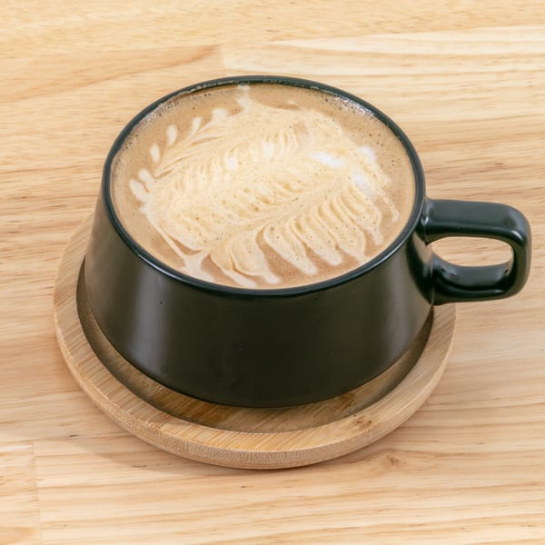 A black mug of latte with intricate foam art sits on a wooden coaster and table.
