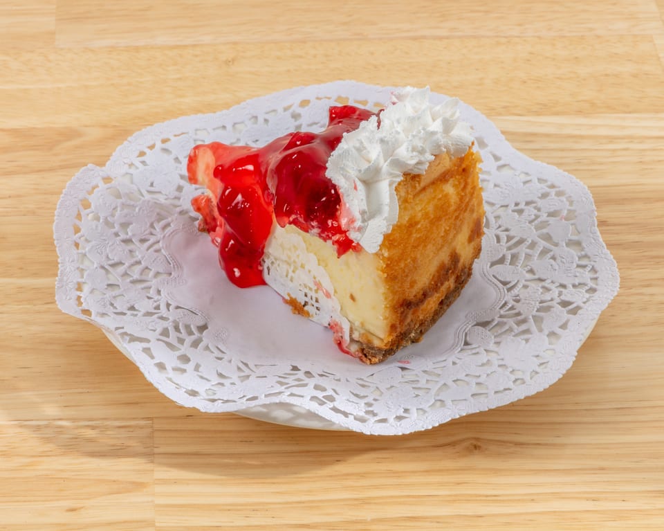 Slice of cheesecake topped with cherry pie filling and whipped cream on a white doily.