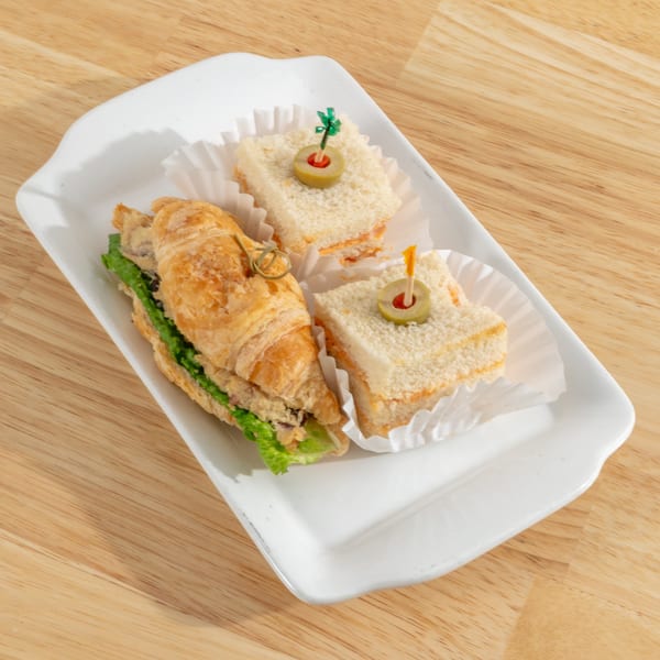 A white rectangular plate holds a croissant sandwich and two small square sandwiches with olive picks.