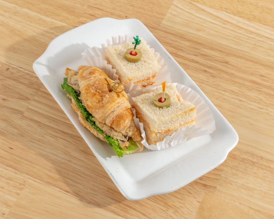 A white rectangular plate holds a croissant sandwich and two small square sandwiches with olive picks.