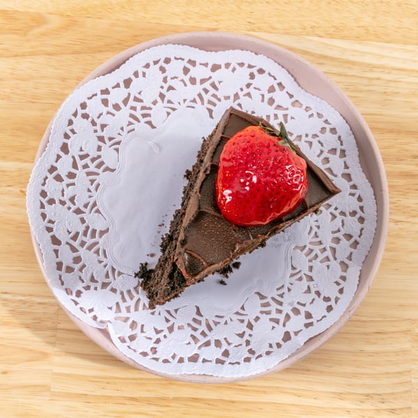 A slice of chocolate cake with a glazed strawberry on top, served on a white doily.