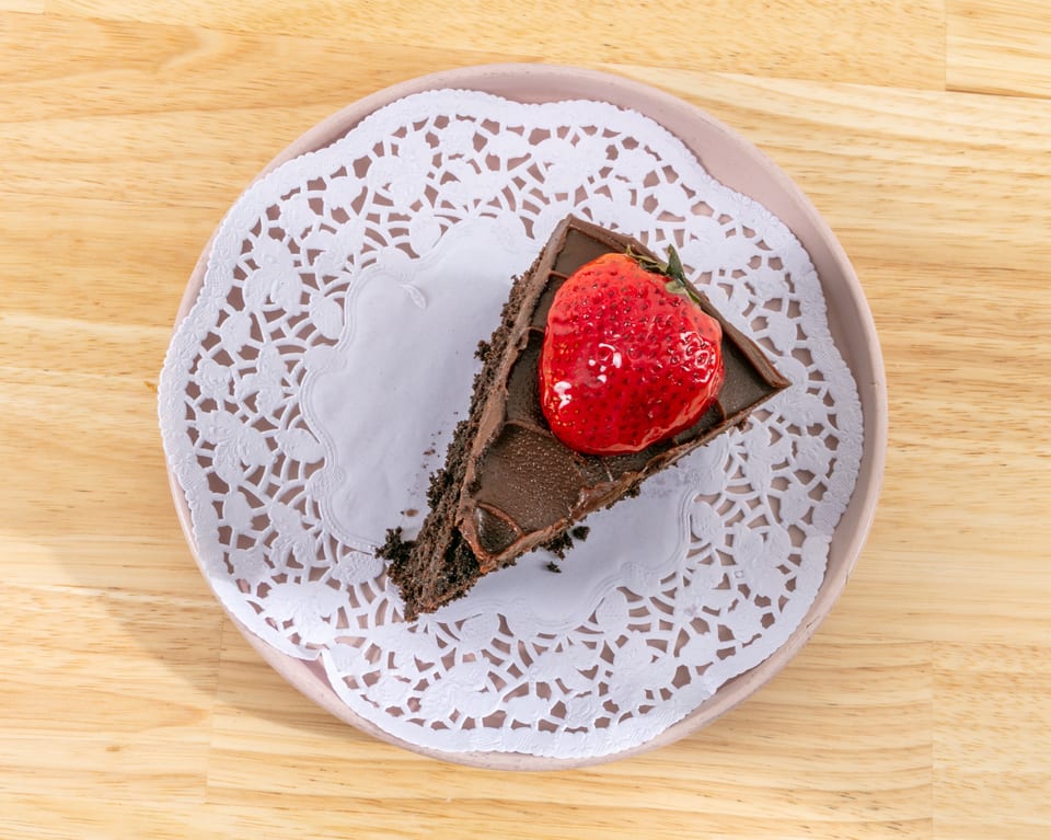 A slice of chocolate cake with a glazed strawberry on top, served on a white doily.